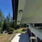 Close-up of a white metal gutter or downspout attachment with two visible screws, shown installed under a house eave with trees and a patio in the background.