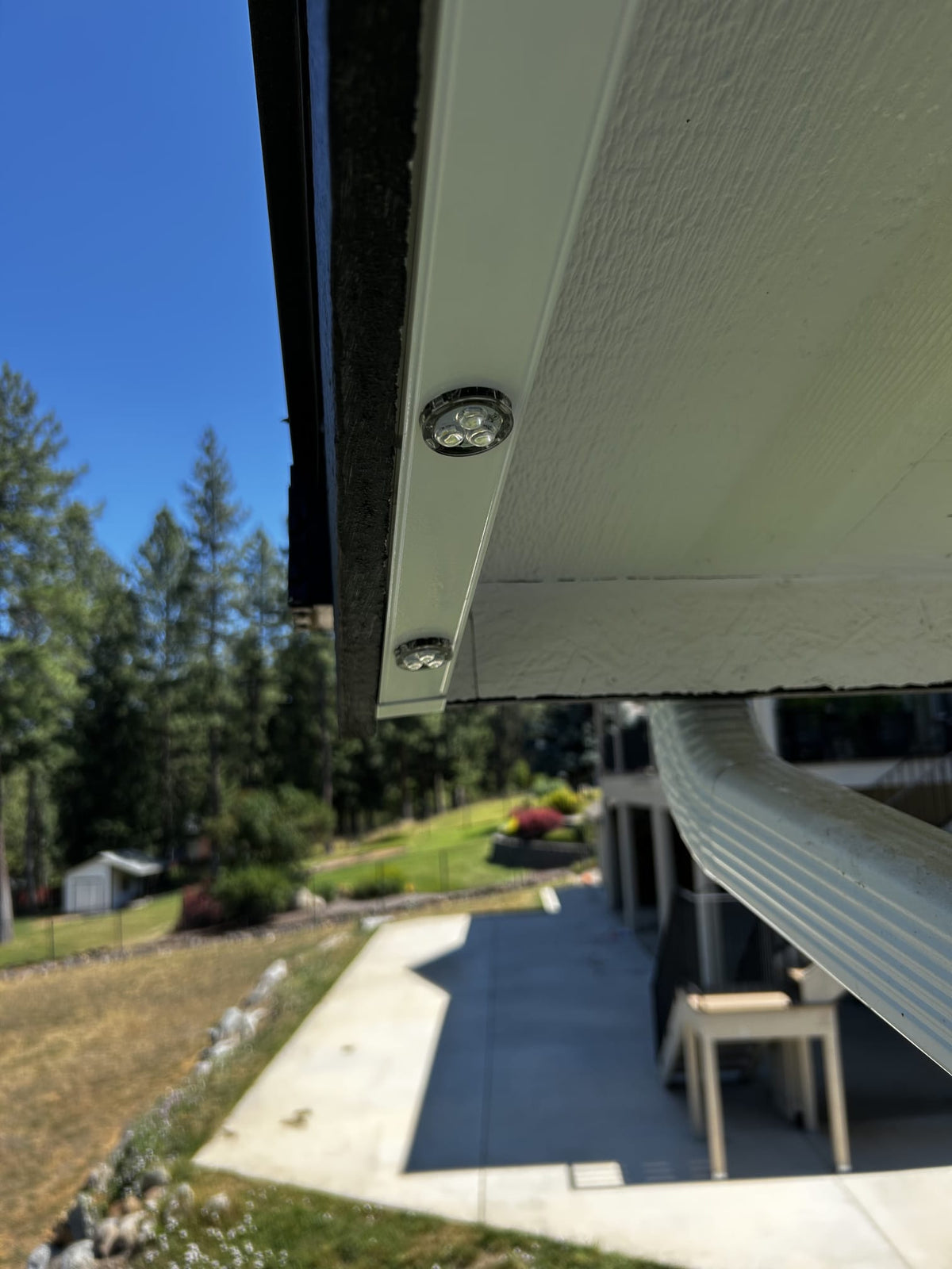 Close-up of a white metal gutter or downspout attachment with two visible screws, shown installed under a house eave with trees and a patio in the background.