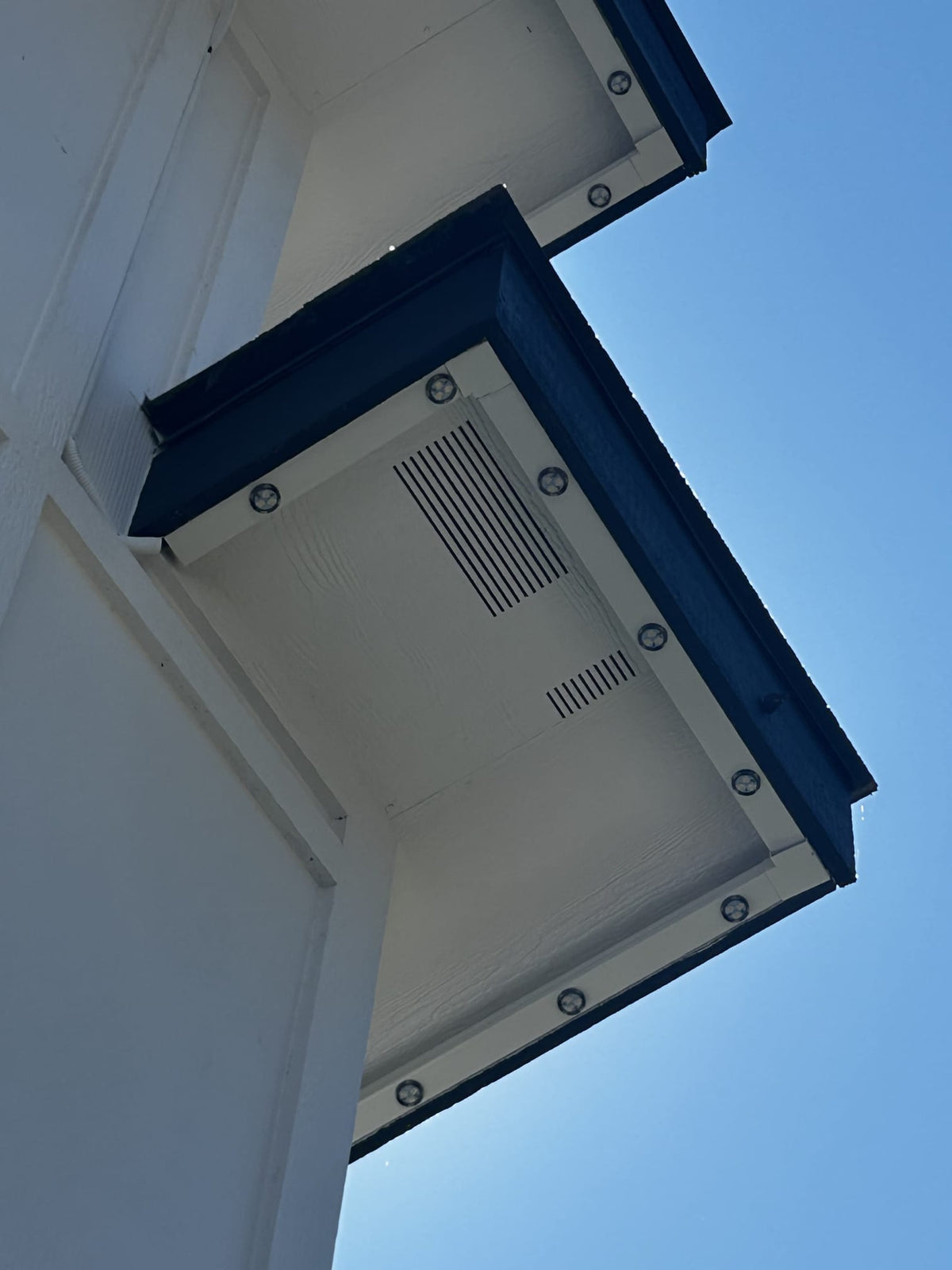An upward-looking view of a white exterior wall corner with dark blue trim and multiple recessed light fixtures, set against a clear blue sky.