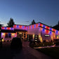 A house at dusk with colorful LED light strips outlining the exterior, casting red, blue, and purple illumination across the walls and roofline. The scene includes a driveway with two dark vehicles, trees, lawn, and small ground lights.