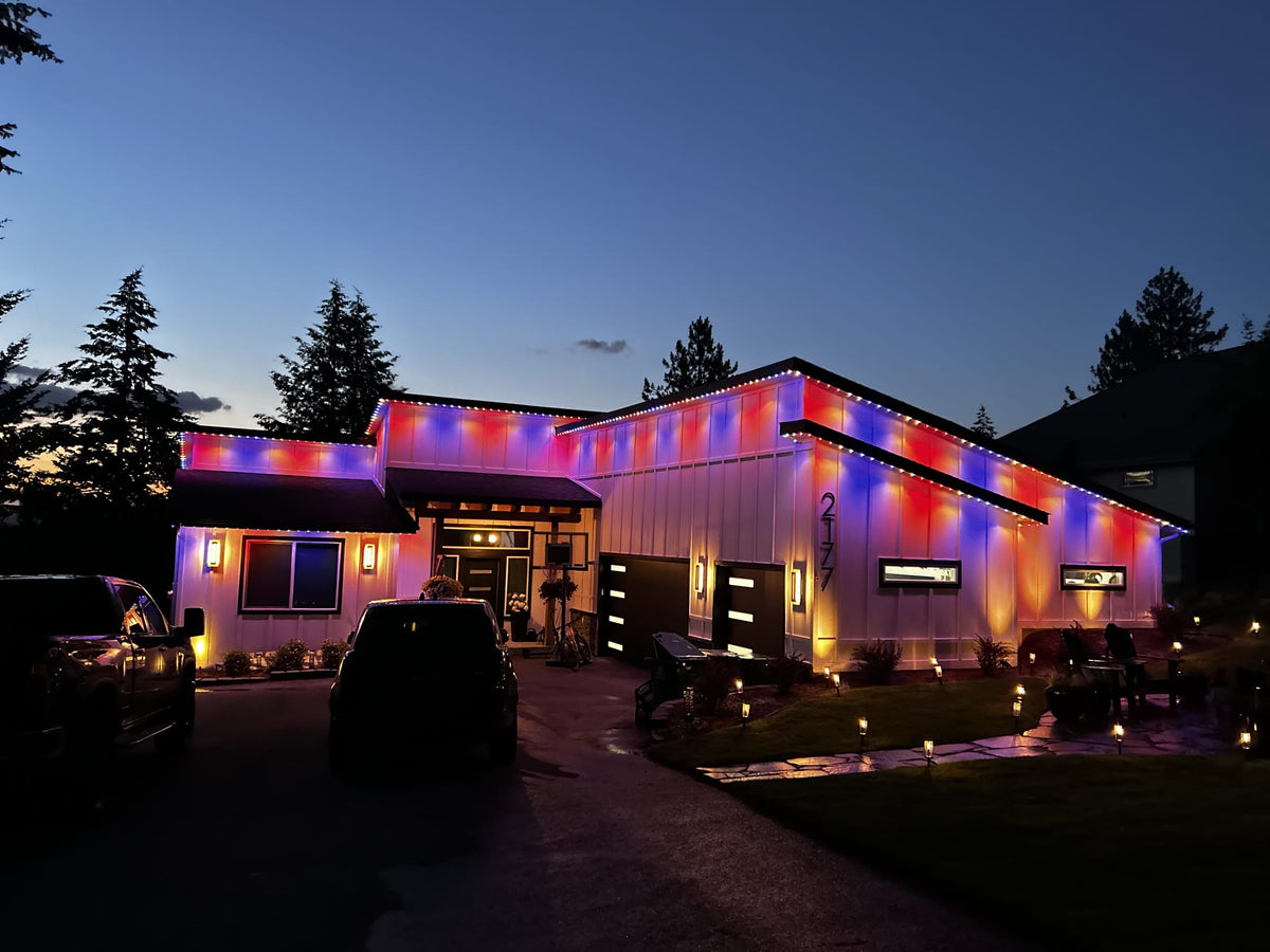 A house at dusk with colorful LED light strips outlining the exterior, casting red, blue, and purple illumination across the walls and roofline. The scene includes a driveway with two dark vehicles, trees, lawn, and small ground lights.